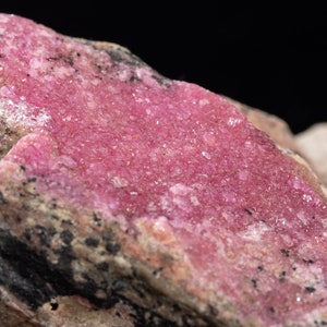 May include: A close-up of a mineral specimen with a vivid pink crystalline formation. The mineral is set against a backdrop of lighter-coloured rock with dark inclusions, creating a striking contrast. The texture is rough and uneven.