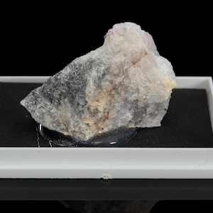 May include: A pale, irregularly shaped mineral specimen, possibly a raw gemstone, sits on a black surface within a white display tray. The mineral has a mix of grey, white, and tan hues, with a slightly rough texture. The background is black.