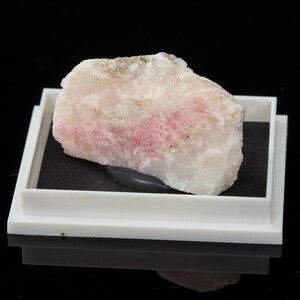 May include: A natural mineral specimen with a pink and white colour gradient. The stone is displayed in a white plastic tray with a black interior, set against a black background. The mineral's texture is coarse and uneven.