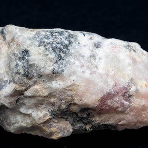 May include: A close-up of a rough, irregularly shaped rock with a mix of colours. The rock has a base of white and light pink, with patches of black and grey throughout. The texture appears coarse and uneven.
