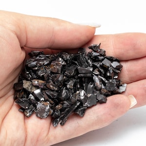 May include: A close-up of a hand holding a collection of dark brown to black, irregularly shaped fragments. The pieces have a reflective, almost metallic appearance. The image highlights the varying sizes and textures of the material.