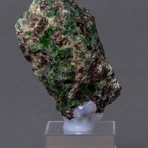 Puede incluir: Un espécimen mineral en bruto con tonos verdes, marrones y rojos. Montado sobre un soporte transparente con una etiqueta que dice "PYROP, CHROMDIOPS Åheim/NO".