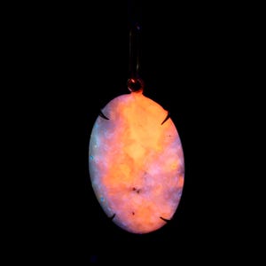 May include: An oval pendant with a vibrant, multicolored design, featuring shades of orange, pink, and blue. The pendant is suspended from a thin, dark-coloured metal loop. The background is solid black, highlighting the pendant's glowing colours.
