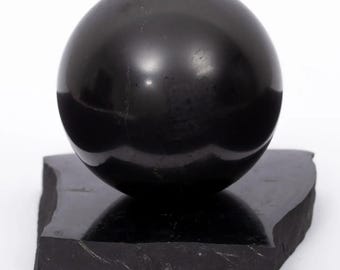 Puede incluir: Escultura de esfera negra pulida sobre una base rectangular oscura. La esfera refleja la luz, creando reflejos. La base tiene un borde rugoso y natural. La escultura está sobre un fondo blanco.
