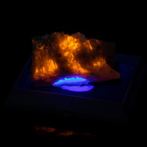 May include: A mineral specimen illuminated with orange and blue light against a black backdrop. The rock has a rough, textured surface, with the light highlighting its crystalline structure. The blue light emanates from a cavity within the rock.