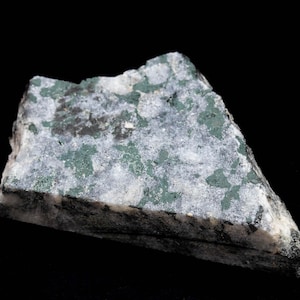 May include: A piece of rock with a rough, uneven surface. It displays a mix of grey, white, and green colours, with darker grey and green patches. The rock is set against a black backdrop.