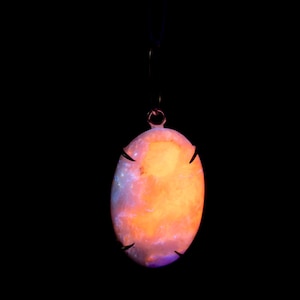 May include: Oval pendant with a glowing orange and purple stone, suspended by a thin black cord. The stone is encased in a dark metal frame with decorative claw-like prongs. The background is solid black.