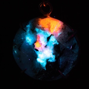 May include: A circular pendant with a dark backdrop, showcasing a luminous display of colours. The pendant features orange, blue, pink, and white hues, creating a striking contrast. It is suspended by a small metal loop.