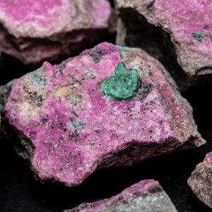 May include: Close-up of several rough-cut rocks with a vibrant pink hue. Some rocks have small patches of green mineral deposits. The rocks have a rough, textured surface and varying shades of pink and grey.