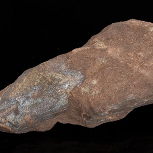 May include: A reddish-brown rock with a rough, textured surface. The rock has a dark, metallic-looking area on one side. The rock is set against a black background.