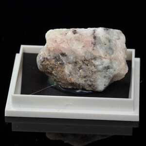 May include: A multi-coloured mineral specimen, predominantly white, pink, grey, and black, with an irregular shape. The rock is presented in a white plastic tray with a black interior, set against a black background.