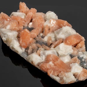 May include: A cluster of peach-colored crystal formations atop a matrix of white and gray minerals. The crystals are elongated and clustered, with a slightly rough texture. The mineral formation is displayed against a dark background.