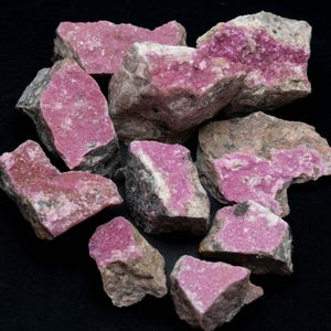 May include: A selection of rough mineral specimens. The rocks are a mix of grey and brown, with vibrant, hot pink crystalline formations throughout. The specimens are irregularly shaped and vary in size, showcasing the raw beauty of the minerals.