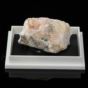 May include: A light pink and beige mineral specimen, possibly a raw gemstone, sits in a white display tray with a black interior. The mineral has a rough, textured surface with dark grey and black inclusions. The background is black.