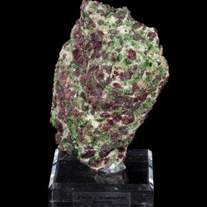 May include: A mineral specimen featuring deep red pyrope garnet crystals embedded in a matrix of vibrant green chromdiopside. The specimen is mounted on a clear acrylic base with a label reading PYROP, CHROMDIOPSID Aheim/NO.
