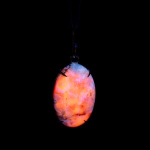 May include: An oval pendant featuring a glowing orange and pink stone, held by a dark metal setting and a thin, dark cord against a black backdrop. The stone has a marbled pattern.