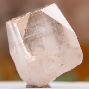 May include: A clear, faceted crystal with a pale peach hue. The crystal has a geometric shape with multiple flat surfaces and a small, reddish-orange inclusion. The base of the crystal is a light gray color.