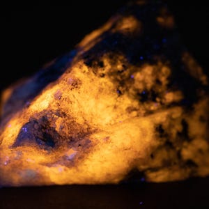 May include: A glowing, irregularly shaped rock with a rough texture. The rock has a vibrant orange and yellow colour, with patches of blue and dark areas. The image is set against a black background, highlighting the rock's luminescence.