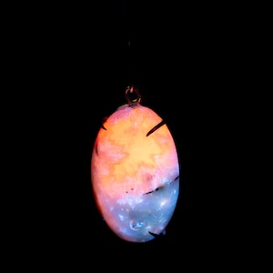 May include: An oval pendant with a copper-coloured loop at the top. The pendant has a glowing, multi-coloured design, with orange and pink hues at the top, transitioning to blue and white at the bottom. Black lines are visible.