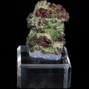 May include: A raw mineral specimen featuring deep red garnet crystals embedded within a matrix of vibrant green epidote. The cluster is mounted on a smooth, light blue base, which is supported by a clear, rectangular acrylic display stand.