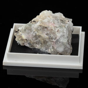 May include: A light grey mineral specimen with pink and black inclusions, presented in a white display tray with a black interior. The rough, crystalline texture of the mineral is visible against a black backdrop. The specimen is irregularly shaped.