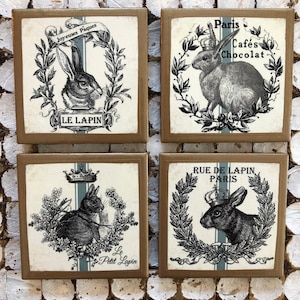 May include: Set of four square ceramic coasters with a vintage design featuring a rabbit with a crown and floral wreath. The coasters have a brown background with white and blue accents. The text on the coasters reads "Joyeuses Pâques", "Le Lapin", "Paris Cafés Chocolat", "Rue de Lapin, Paris", and "Le Petit Lapin".