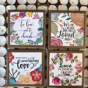 May include: Four square wooden coasters with gold trim and floral designs. Each coaster features a different inspirational quote about love. The quotes are: "To love and to cherish", "We love because He first loved us 1 John 4:19", "I have loved you with an everlasting love, Jeremiah 31:3", and "Love one another like I have loved you, John 13:34".