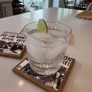 Op de afbeelding: Een helder glas gevuld met ijsblokjes en een partje limoen, rustend op een vierkante onderzetter met een zwart-wit krantenprint met de tekst YOU CAN DRINK REPEAL VOTED. De onderzetter heeft een bruine rand en een glanzende afwerking.
