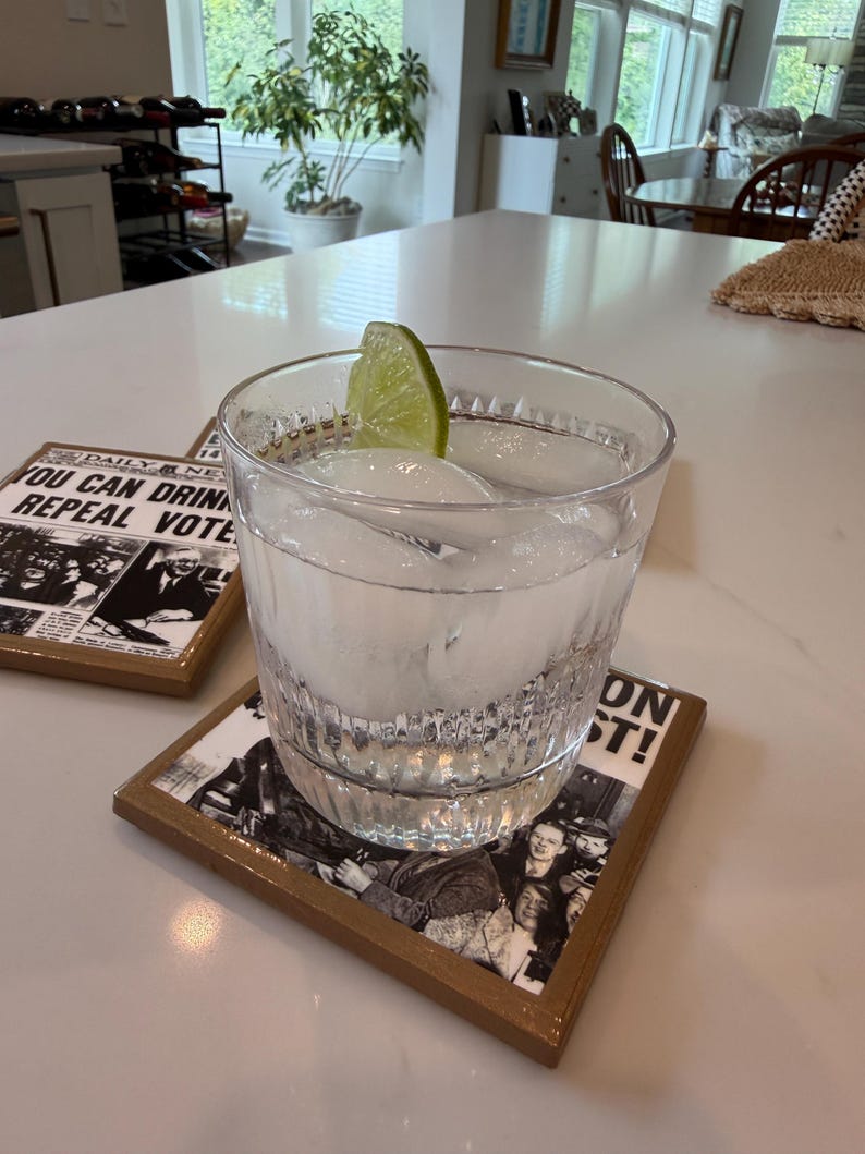 Op de afbeelding: Een helder glas gevuld met ijsblokjes en een partje limoen, rustend op een vierkante onderzetter met een zwart-wit krantenprint met de tekst YOU CAN DRINK REPEAL VOTED. De onderzetter heeft een bruine rand en een glanzende afwerking.
