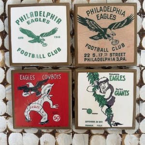 May include: Four square Philadelphia Eagles football club signs. Each sign features the team's logo and text. The signs have a brown border and are set against a white and brown background. The signs include the text "Eagles Cowboys" and "New York Giants".