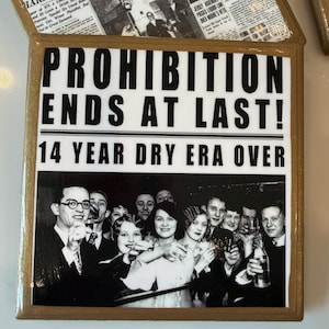 Op de afbeelding: Een vierkante decoratieve tegel met een goudkleurige rand en een zwart-witte afbeelding in krantenstijl. De tekst luidt PROHIBITION ENDS AT LAST! 14 YEAR DRY ERA OVER, vergezeld van een vintage foto van een groep mensen die aan het vieren zijn.