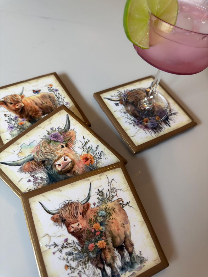 Puede incluir: Tres posavasos cuadrados con bordes dorados, cada uno con una ilustraci&oacute;n impresa de una vaca Highland marr&oacute;n adornada con flores coloridas. Un posavasos sostiene una copa con una bebida rosa y una rodaja de lima.