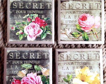May include: Four square coasters with a rustic woodgrain background and floral designs. Each coaster features the word "SECRET" and the French phrase "POUR TEINDRE LA FLEUR D'IMMORTELE" in gold lettering. The floral designs include pink roses, butterflies, and a yellow flower.