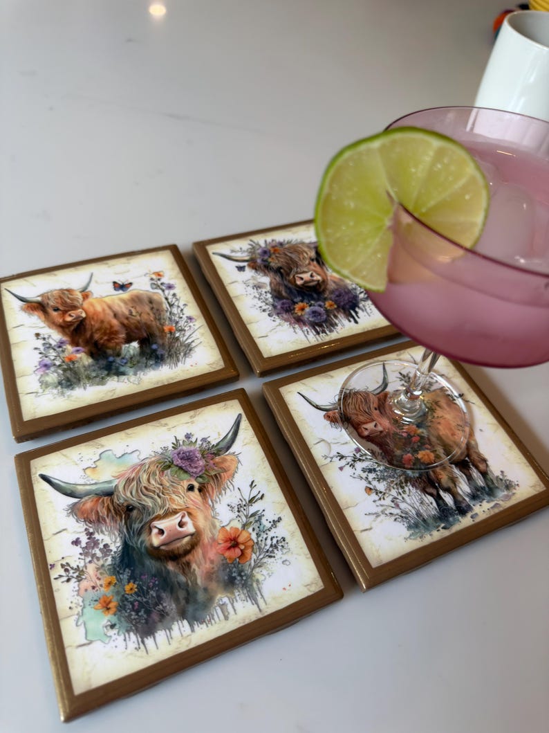 Puede incluir: Cuatro posavasos cuadrados con una ilustraci&oacute;n en acuarela de una vaca Highland con detalles florales. Un posavasos est&aacute; parcialmente cubierto por una copa con un l&iacute;quido rosa y una rodaja de lima. Los posavasos tienen un borde marr&oacute;n.