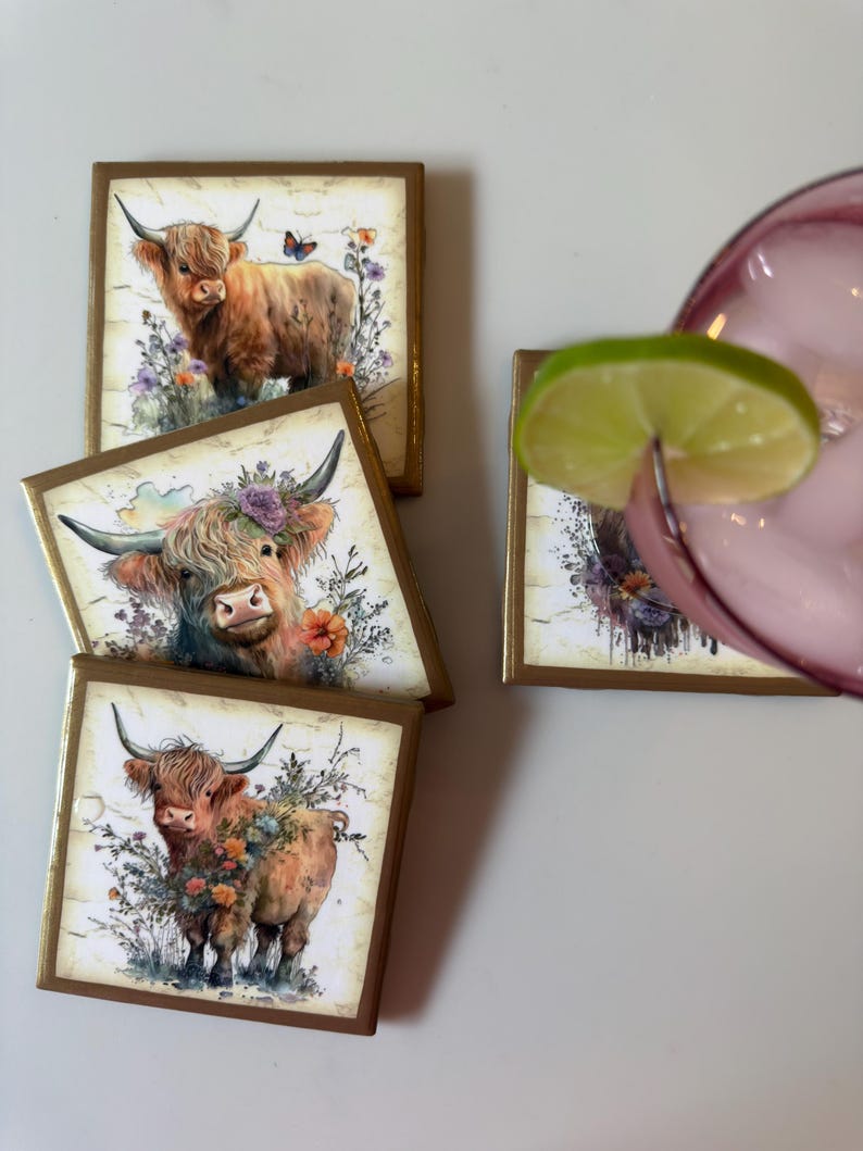 Puede incluir: Cuatro posavasos cuadrados con bordes dorados, cada uno con una ilustraci&oacute;n estilo acuarela de una vaca Highland rodeada de coloridas flores silvestres y vegetaci&oacute;n. Un posavasos est&aacute; parcialmente oculto por un vaso con una rodaja de lima.