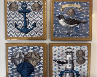 May include: A set of four square coasters with gold-toned borders. The designs feature nautical themes including a blue anchor, a seagull with the text GULL, a collection of seashells, and a lighthouse with the text SEASIDE on a blue background.