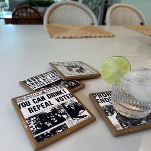 Op de afbeelding: Een set vierkante onderzetters met zwart-wit krantenknipsels over het einde van de drooglegging. De tekst luidt "YOU CAN DRINK! REPEAL VOTED" en "PROHIBITION ENDS AT LAST". Een glas water met een schijfje limoen staat op een van de onderzetters.