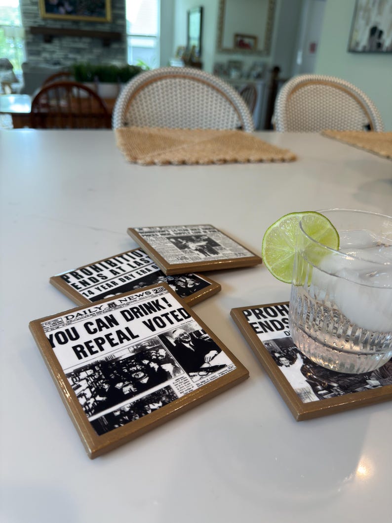 Op de afbeelding: Een set vierkante onderzetters met zwart-wit krantenknipsels over het einde van de drooglegging. De tekst luidt "YOU CAN DRINK! REPEAL VOTED" en "PROHIBITION ENDS AT LAST". Een glas water met een schijfje limoen staat op een van de onderzetters.