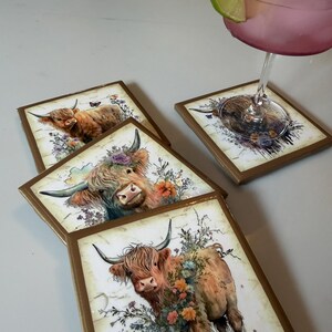 Puede incluir: Un juego de tres posavasos cuadrados con bordes dorados, cada uno con una ilustraci&oacute;n impresa de una vaca Highland adornada con coloridos acentos florales. Un posavasos sostiene una copa con l&iacute;quido rosa y una rodaja de lima.