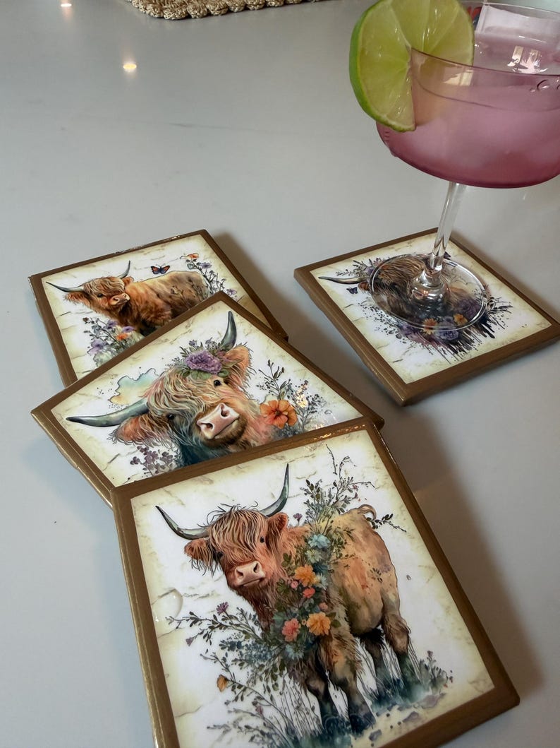 Puede incluir: Un juego de tres posavasos cuadrados con bordes dorados, cada uno con una ilustraci&oacute;n impresa de una vaca Highland adornada con coloridos acentos florales. Un posavasos sostiene una copa con l&iacute;quido rosa y una rodaja de lima.