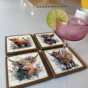 Puede incluir: Cuatro posavasos cuadrados con bordes marrones que presentan una ilustraci&oacute;n en acuarela de una vaca Highland adornada con flores. Una copa con un l&iacute;quido rosa, cubitos de hielo y una rodaja de lima descansa sobre uno de los posavasos.