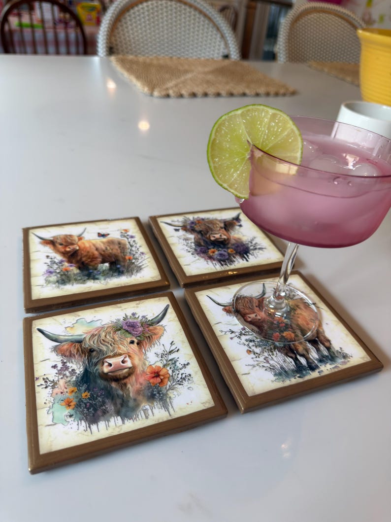 Puede incluir: Cuatro posavasos cuadrados con bordes marrones que presentan una ilustraci&oacute;n en acuarela de una vaca Highland adornada con flores. Una copa con un l&iacute;quido rosa, cubitos de hielo y una rodaja de lima descansa sobre uno de los posavasos.