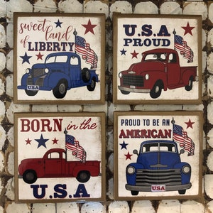 May include: Four wooden signs with patriotic themes. Each sign features a vintage pickup truck with an American flag and text. The signs read "Sweet land of Liberty", "U.S.A Proud", "Born in the U.S.A", and "Proud to be an American".