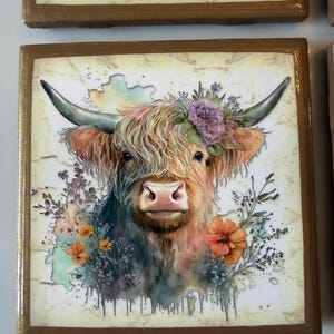 Puede incluir: Un azulejo decorativo cuadrado con una ilustraci&oacute;n estilo acuarela de una vaca Highland con pelaje largo y desgre&ntilde;ado, cuernos curvados y una corona de flores moradas y naranjas. El fondo tiene una textura color crema tipo m&aacute;rmol con gotas.