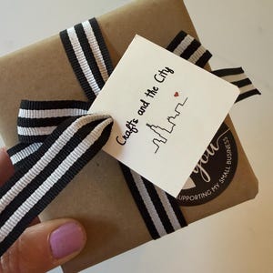 Puede incluir: Un paquete cuadrado envuelto en papel marr&oacute;n, atado con una cinta a rayas blancas y negras. Se adjunta una etiqueta cuadrada blanca con el texto "Crafts and the City", una ilustraci&oacute;n de un horizonte y un peque&ntilde;o coraz&oacute;n rojo, y una pegatina.