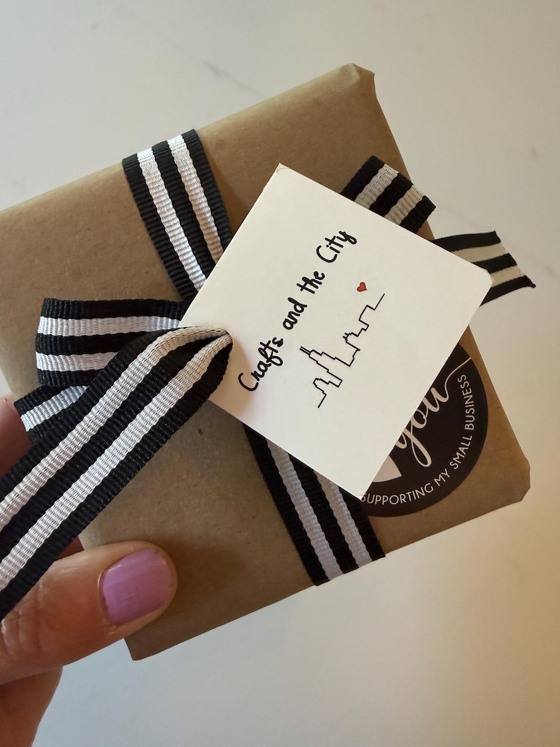 Puede incluir: Un paquete cuadrado envuelto en papel marr&oacute;n, atado con una cinta a rayas blancas y negras. Se adjunta una etiqueta cuadrada blanca con el texto "Crafts and the City", una ilustraci&oacute;n de un horizonte y un peque&ntilde;o coraz&oacute;n rojo, y una pegatina.