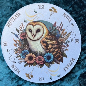 May include: Round divination board featuring a detailed illustration of a barn owl surrounded by flowers. The board has text that reads "Yes", "Maybe", "No", and "Rephrase". The owl has brown and white feathers, and the flowers are in shades of blue, pink, and orange.