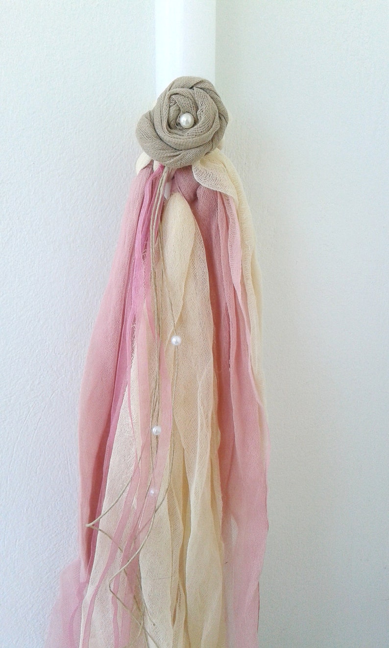 May include: A white candle with a pink and cream fabric decoration. The fabric is tied with a brown ribbon and has a small pearl embellishment.