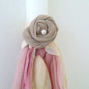 May include: A white candle decorated with a beige fabric rose and pearl embellishment. The candle is adorned with pink and cream fabric ribbons.