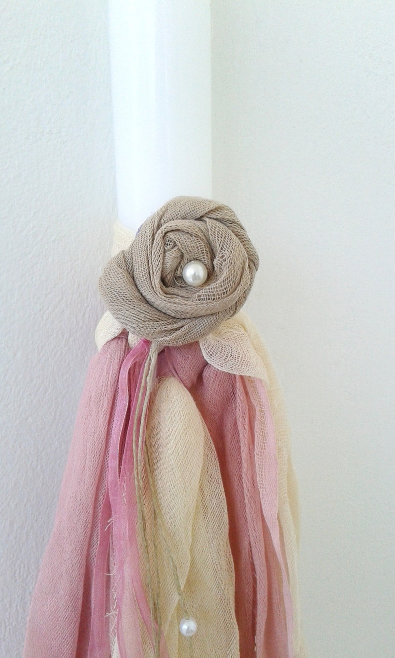 May include: A white candle decorated with a beige fabric rose and pearl embellishment. The candle is adorned with pink and cream fabric ribbons.